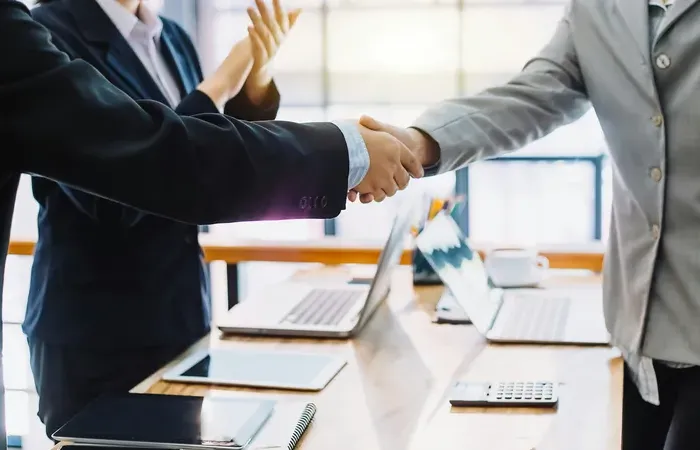 Two professionals shaking hands across a wooden desk with laptops and documents during a business meeting on Town Service Spot